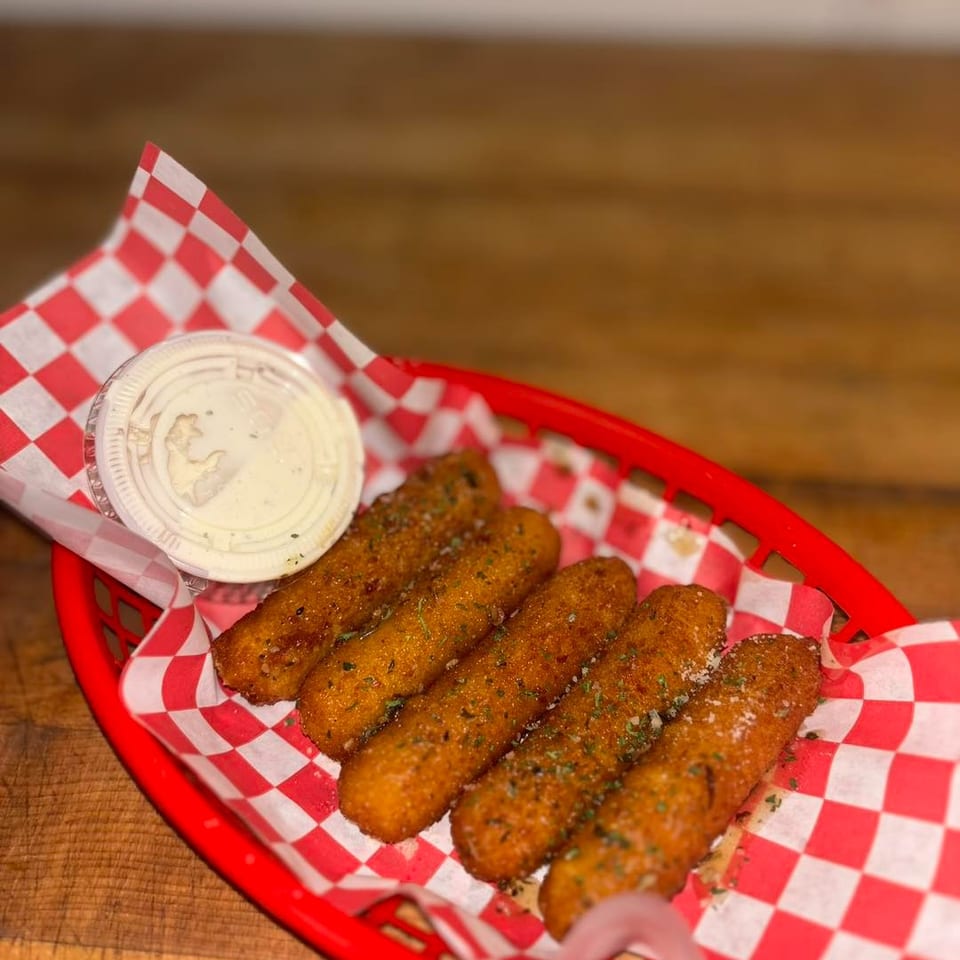 Five golden-brown mozzarella sticks with herbs and a side of ranch in a red basket with checkered paper.