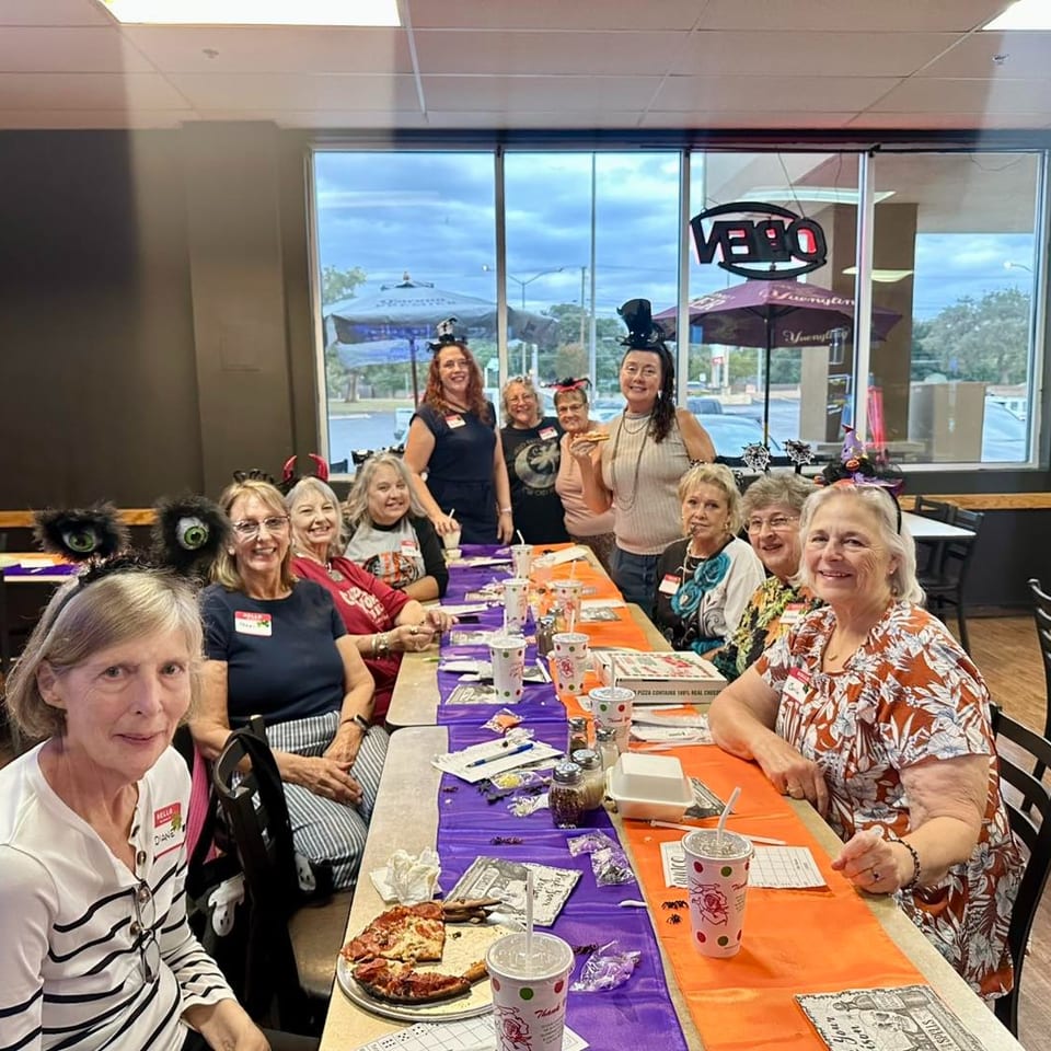 A group of women in Halloween headbands and costumes sit at a long table with pizza and drinks.