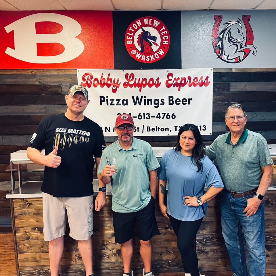 Four people stand in front of a Bobby Lupos Express sign, with a wooden wall behind them.