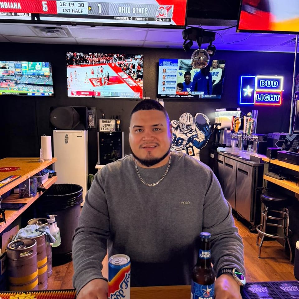 Man sitting at a bar with drinks, surrounded by multiple TVs showing sports games.