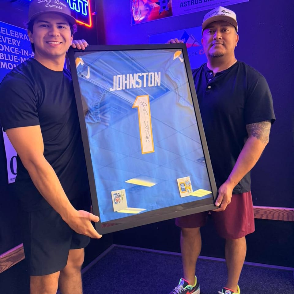 Two men holding a framed blue sports jersey with "JOHNSTON" and the number "1" on it.
