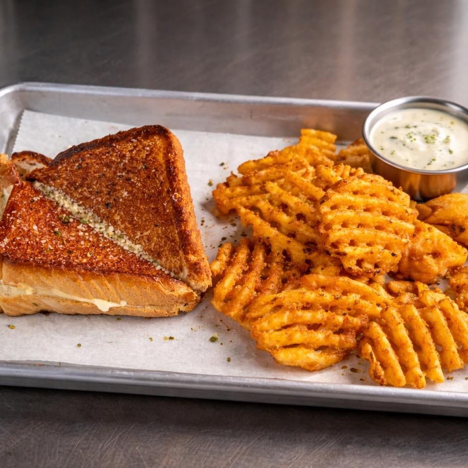 Grilled cheese sandwich with waffle fries and a side of dipping sauce on a metal tray.